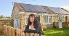 A woman leaning over her garden fence, smiling at the camera with her house, that has had energy effiiceny upgrades, in the background