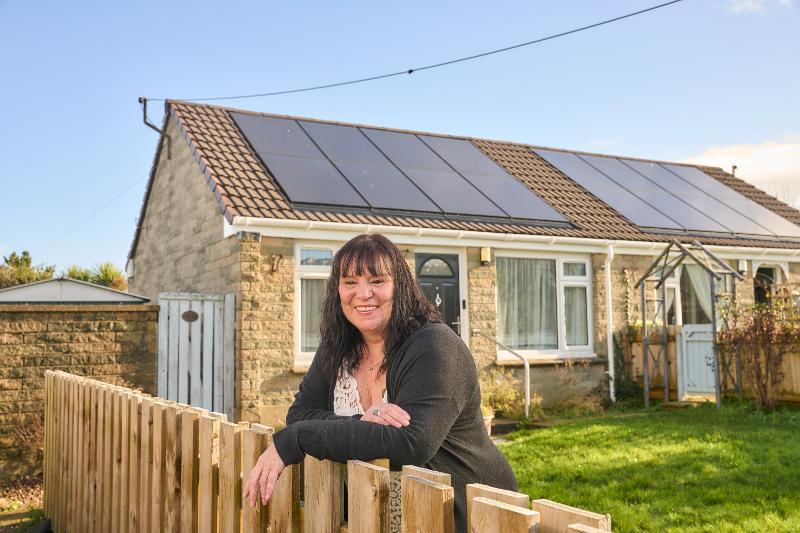 A woman leaning over her garden fence, smiling at the camera with her house, that has had energy effiiceny upgrades, in the background