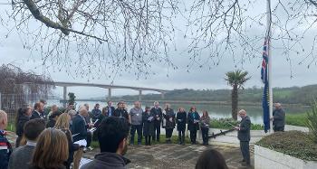 A group gathers outside Riverbank House beside the River Torridge for a Commonwealth Flag‑raising event. The crowd forms a semicircle near a flagpole while the flag is raised. The Torridge Bridge spans the river in the background under cloudy skies.
